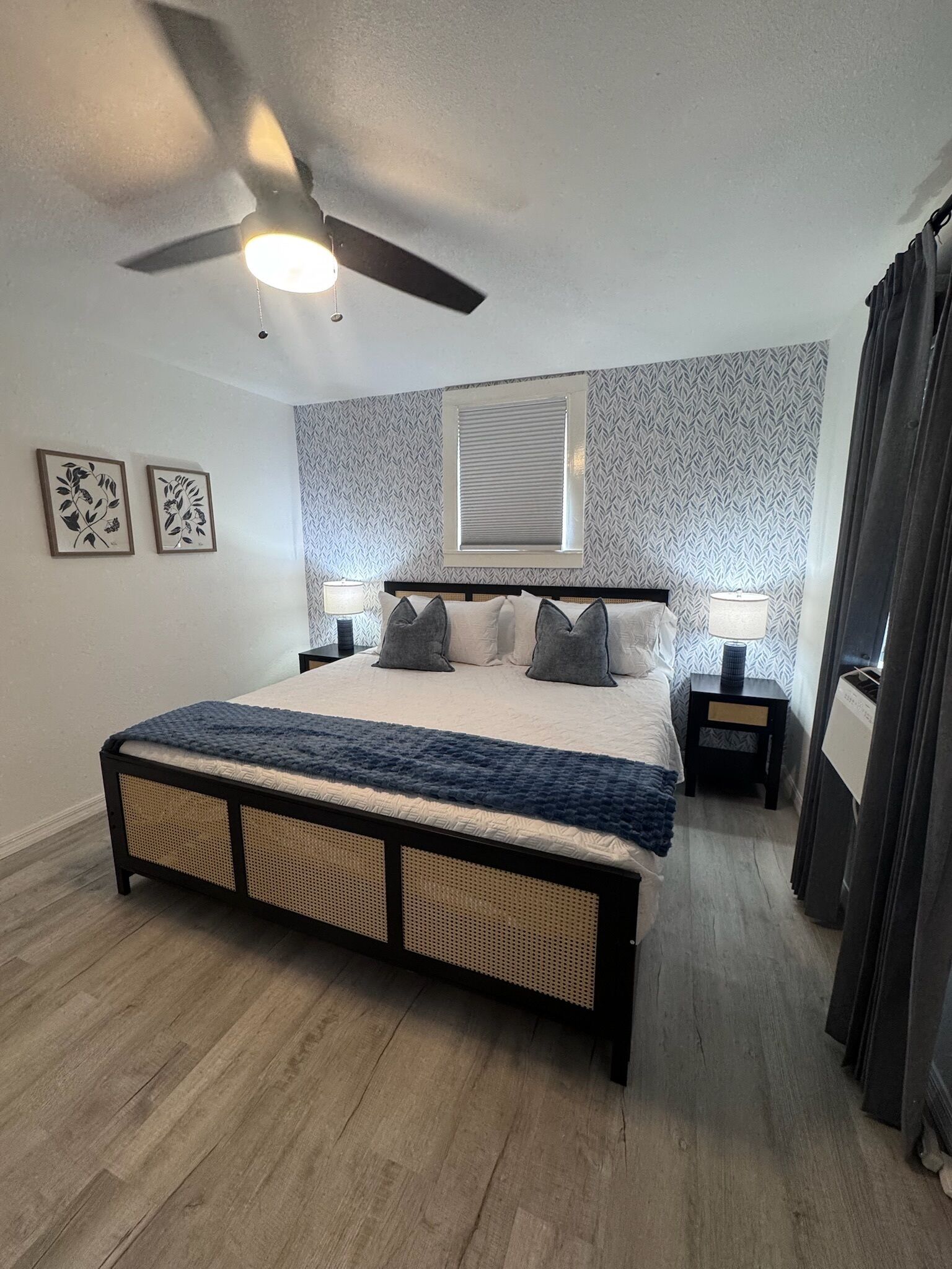 Un dormitorio con cama, dos mesitas de noche, ventilador de techo y ventana con persianas.
