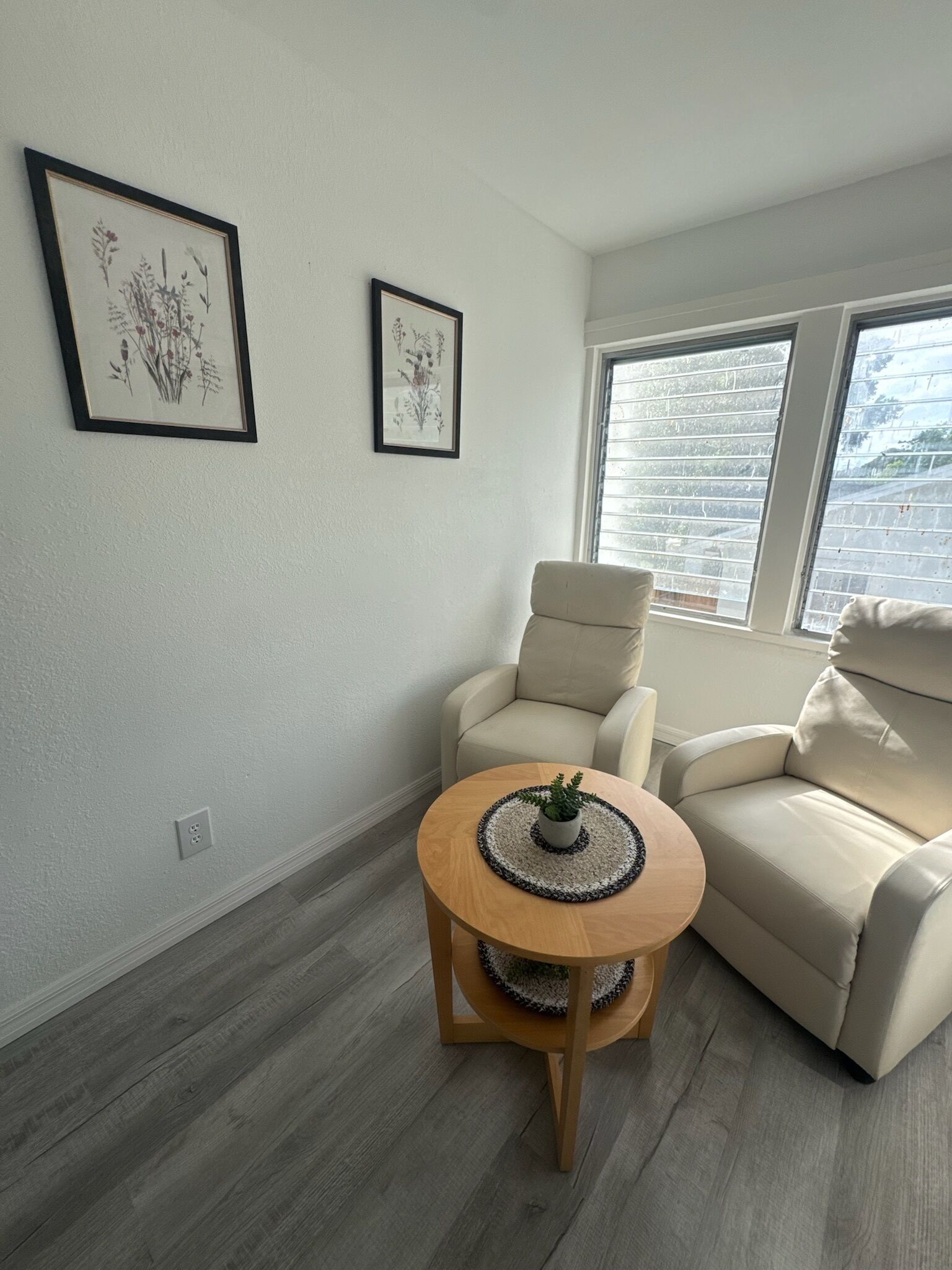 A living room with a beige sofa, a wooden coffee table with a plant, and two framed botanical prints on the wall.