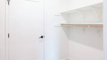 A room with a white door, beige carpet, and white walls. There are two white shelves with hooks on the wall.