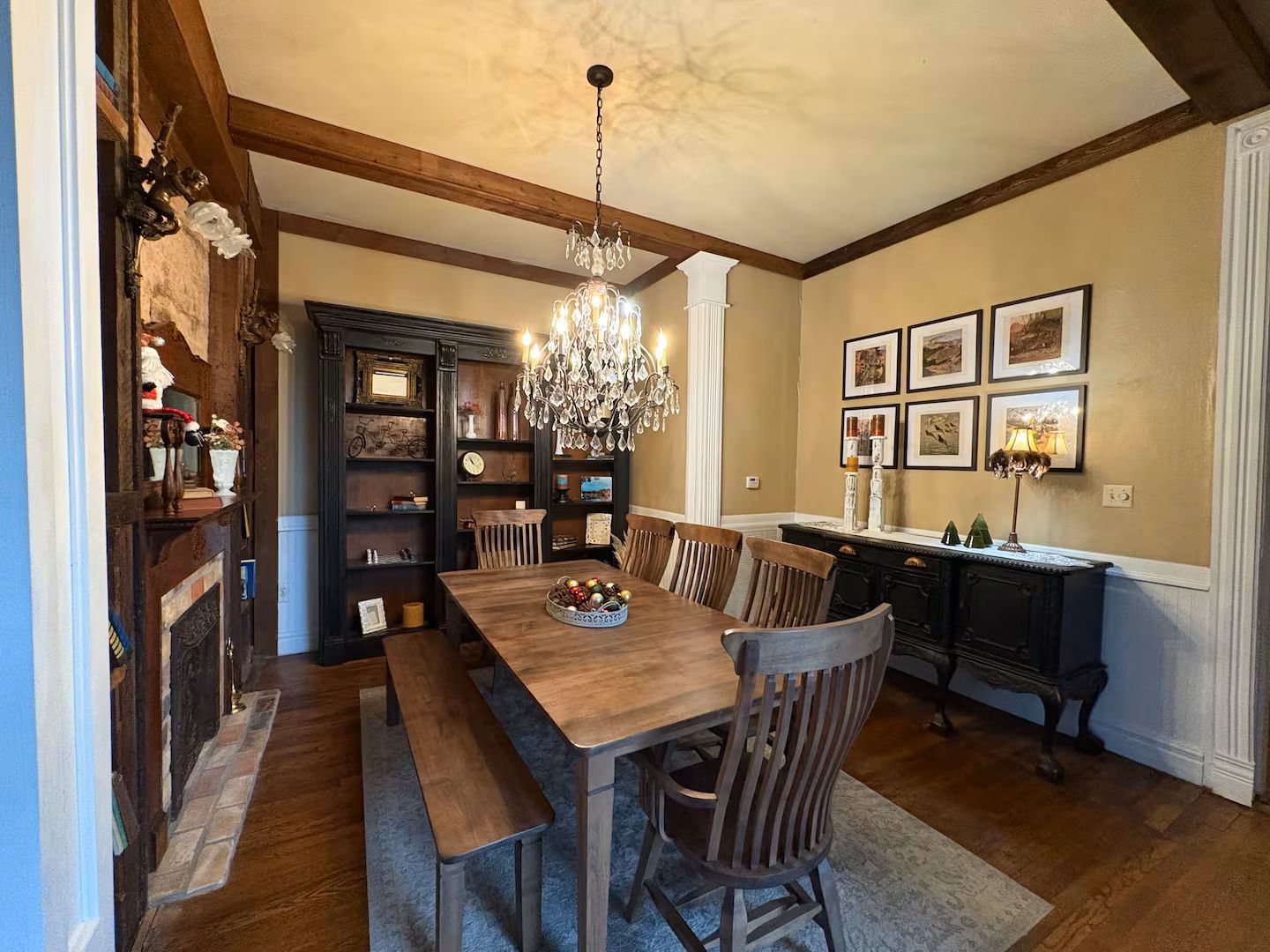 A dining area with a wooden table, benches, and a chandelier. There are bookshelves and framed pictures on the walls.