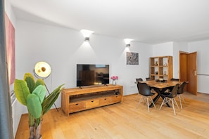 A modern living room with a wooden entertainment unit, a flat-screen TV, a dining table with chairs, and a potted plant. - Spacious vacation rental with mountain views and a terrace near the highway and Lake Simssee (Rohrdorf)