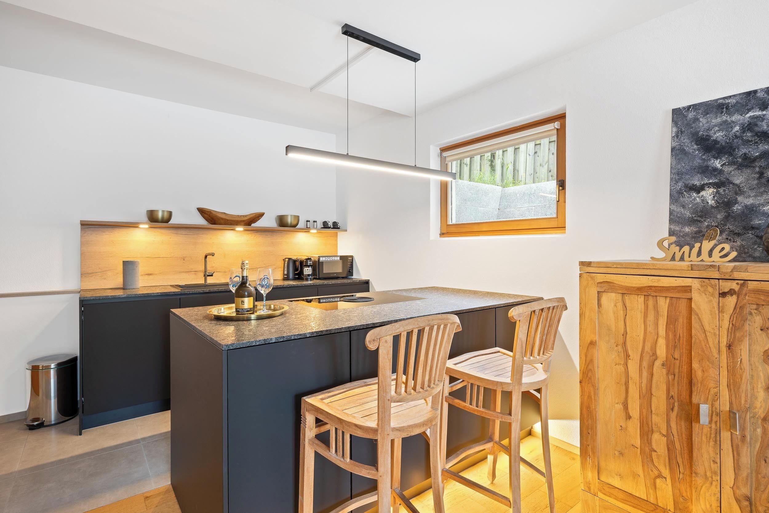 A modern kitchen with a central island, bar stools, a wooden cabinet, and a view of the outdoors.