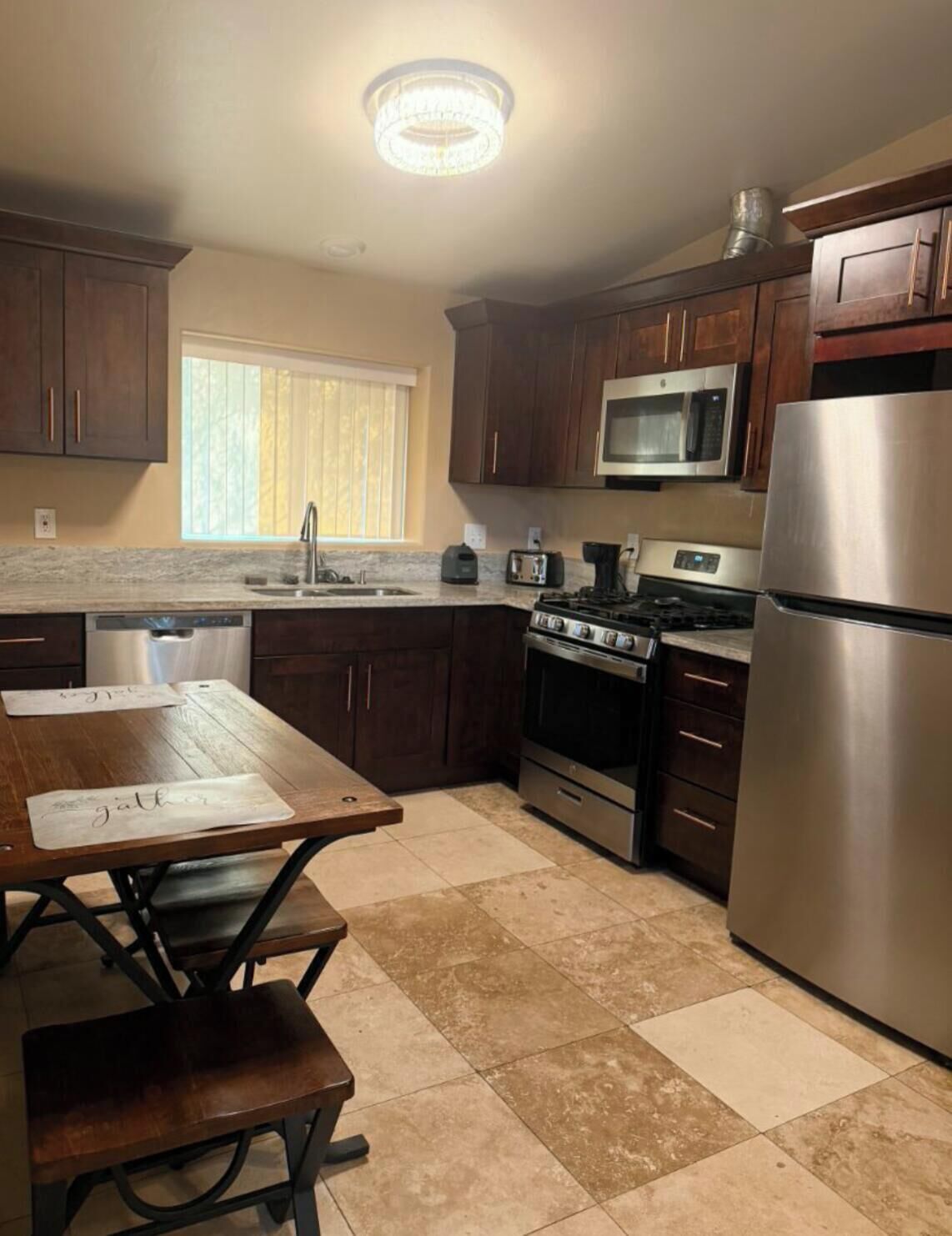 A kitchen with dark wooden cabinets, stainless steel appliances, a window with blinds, and a table with a placemat.