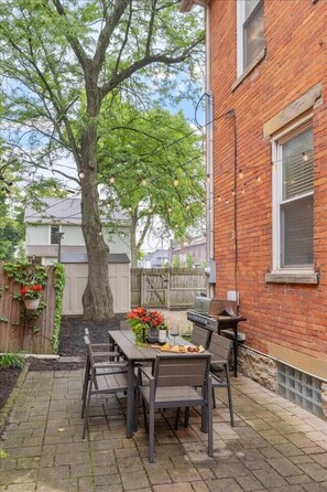 Ein Backsteinhaus mit einer Terrasse, ausgestattet mit Tisch, Stühlen, einem Grill und einem großen Baum.