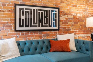 Un sofá azul con detalles capitonados frente a una pared de ladrillo, con un cartel enmarcado encima que dice 'COLUMBIA'.