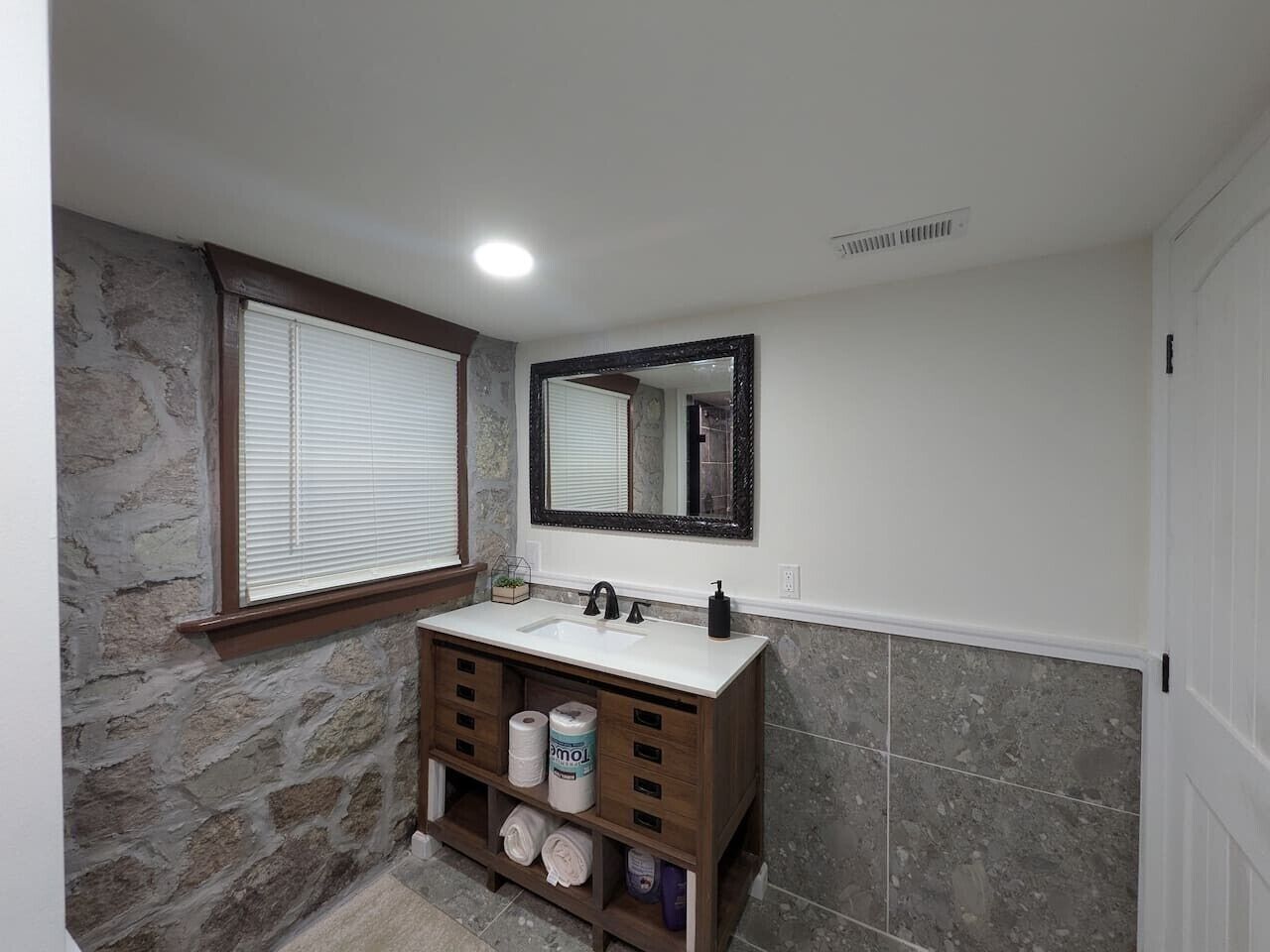 A bathroom with a stone wall, a wooden vanity, a sink, a mirror, and a window with blinds.