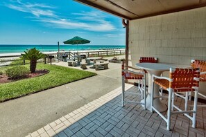 An outdoor patio with a view of the beach, featuring tables and chairs, a small palm tree, and a green umbrella. - Polynesian #108 (Panama City Beach)