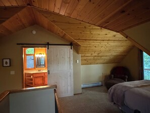 A bedroom with a sloped ceiling, a bed, a chair, and a bathroom with a window.