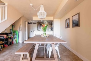 Un coin repas avec une table en bois, des chaises blanches, un lustre et une vue sur la cuisine.