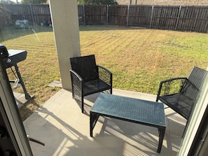 Eine Terrasse mit zwei Stühlen und einem Tisch, einem Betonboden und einem grünen Rasen mit einem Holenzaun im Hintergrund.