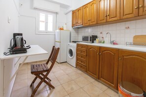 A compact kitchen with wooden cabinets, a washing machine, a microwave, and a dining area with a chair. -  Live Like a Local, Steps From Acropolis | Homey, Roomy & Sleeps 4 (Athina)