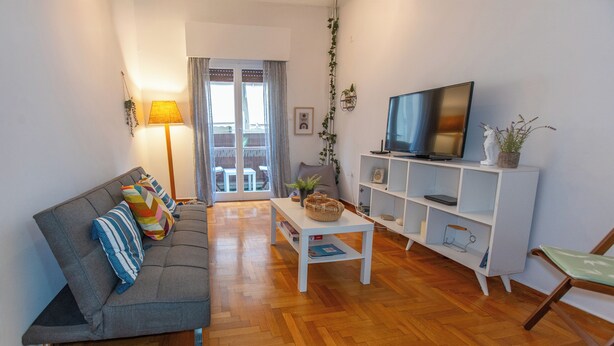 A living room with a sofa, a coffee table, a TV on a stand, and a view through a glass door to an outdoor area. -  Live Like a Local, Steps From Acropolis | Homey, Roomy & Sleeps 4 (Athina)