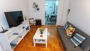 A living room with a flat-screen TV, a white coffee table with a plant, a gray sofa with colorful cushions, and a wooden herringbone floor. -  Live Like a Local, Steps From Acropolis | Homey, Roomy & Sleeps 4 (Athina)