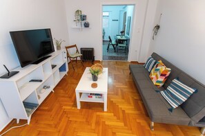 A living room with a flat-screen TV, a white coffee table with a plant, a gray sofa with colorful cushions, and a wooden herringbone floor. -  Live Like a Local, Steps From Acropolis | Homey, Roomy & Sleeps 4 (Athina)