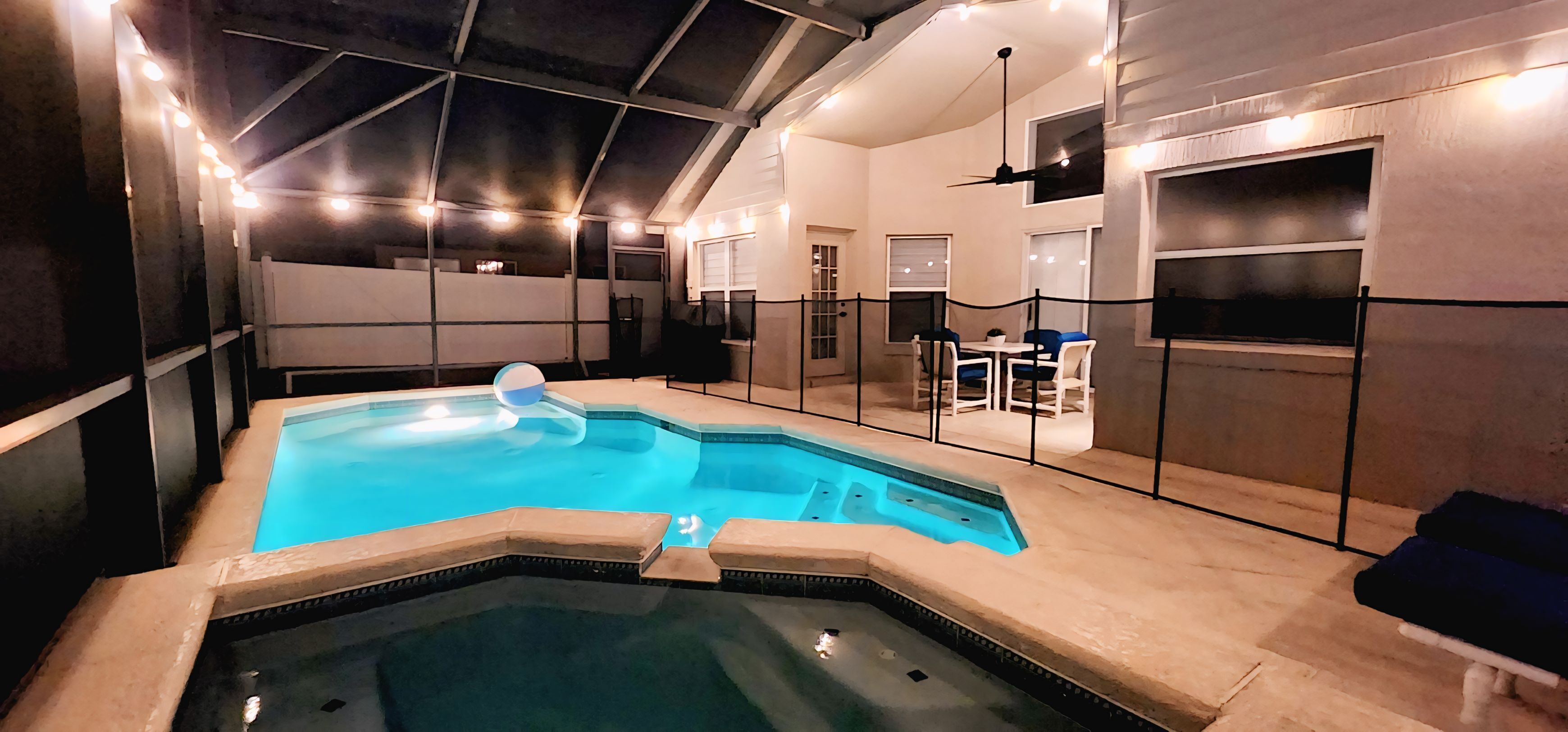 Una piscina interior con hidromasaje, rodeada por una cerca y un área de descanso con mesas y sillas.