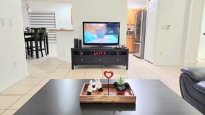A modern living room with a flat-screen TV, a dark-colored sofa, a coffee table with a heart-shaped decoration, and a wooden cabinet with the word 'LOVE' on it. - Lovely home, Quiet Escape Near Disney (Kissimmee)