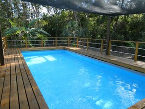 A rectangular pool with a wooden deck and a safety railing, surrounded by lush greenery.