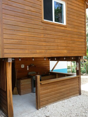A wooden cabin with a kitchen area, a window, and a view of the outdoors. - Authentic Sarteneja– Pool, Birds & Tranquility (Sarteneja)