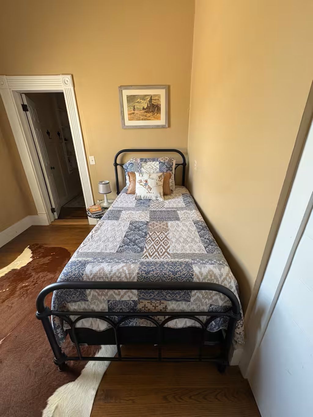 A narrow room with a bed, a door, and a painting on the wall.
