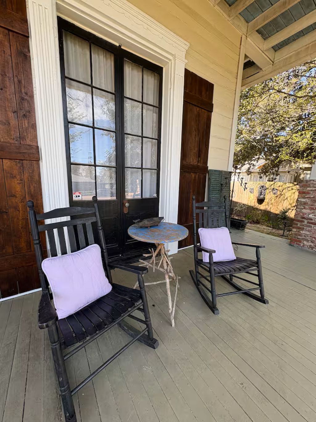A porch with two rocking chairs and a small table, wooden shutters, and a brick wall.