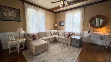 A living room with a sectional sofa, a small table with a lamp, a decorative mirror, and a ceiling fan.