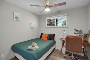 A bedroom with a bed, a desk, a chair, a ceiling fan, and a window.