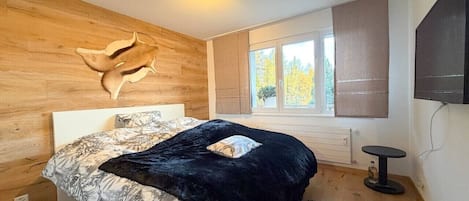 Un dormitorio con cama, cabecera de madera, ventana con persianas, televisor y una mesita redonda.