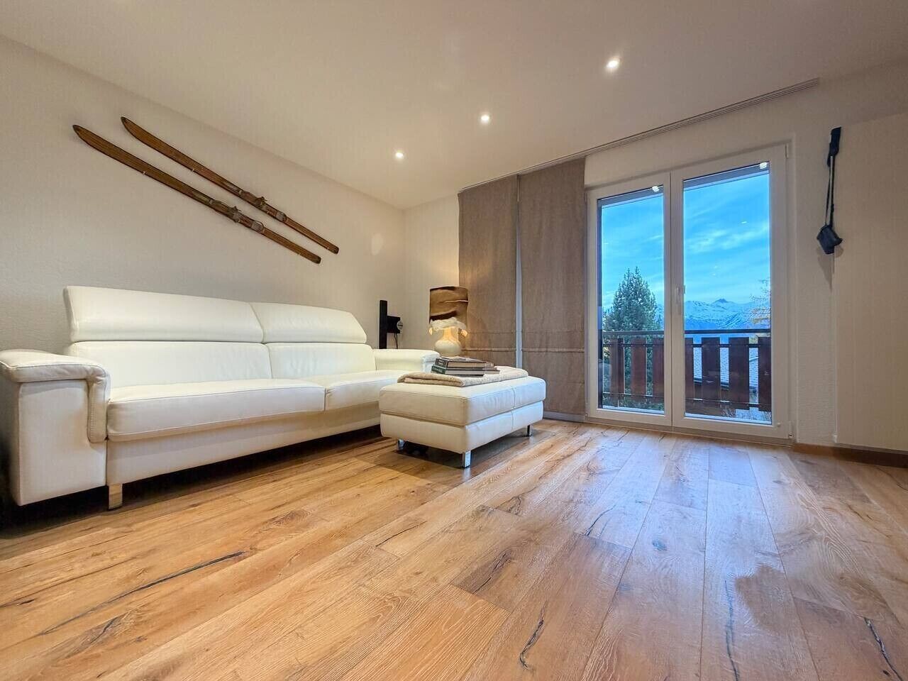 Un salon spacieux avec un canapé blanc, un petit pouf et une grande fenêtre donnant sur l’extérieur.