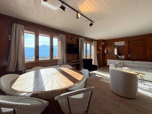 Ein geräumiges Zimmer mit einem runden Holztisch, umgeben von Stühlen, einer Couch und einem Fernseher. Große Fenster bieten Blick auf die Berge.