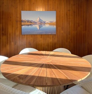 Ein runder Holztisch mit einer eingekerbten Mitte, umgeben von weiß gepolsterten Stühlen, vor einer holzverkleideten Wand mit einem gerahmten Landschaftsbild darüber.