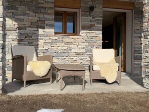 A stone building with a patio area featuring two wicker chairs and a small table. - A fully equipped chalet with a magnificent view of the Alps! (Crans-Montana)