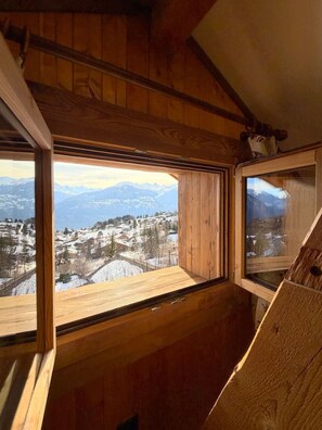 A wooden interior with a large window offering a view of a snowy landscape and mountains. - A fully equipped chalet with a magnificent view of the Alps! (Crans-Montana)