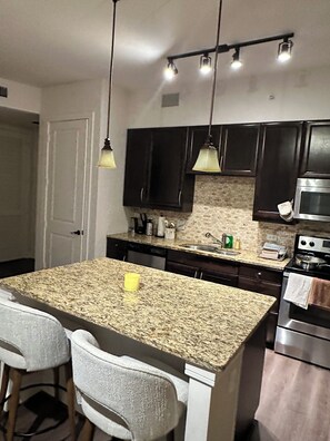 A kitchen with a granite countertop, dark cabinets, stainless steel appliances, and pendant lighting. - Cozy 1-bedroom apartment with backyard in Postoak/ Galleria Houston (Houston)