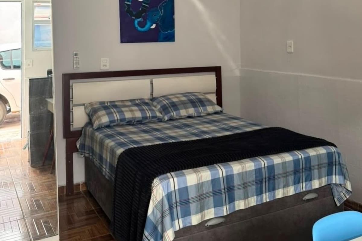 A bedroom with a bed, a blue and white checkered blanket, a dark headboard, and a painting of a blue elephant on the wall.