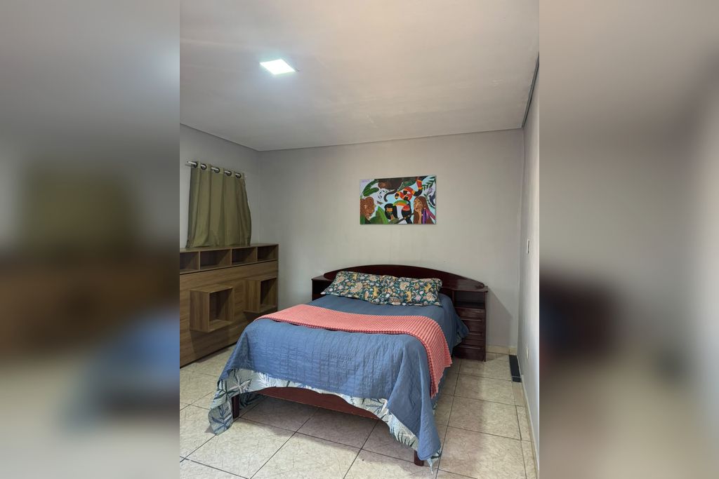 Une chambre à coucher avec un lit, une commode en bois, un tableau accroché au mur et un rideau.