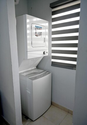 Ein kleiner Waschraum mit einer Frontlader-Waschmaschine und einem Trockner, begrenzt von einer weißen Wand mit einem Fenster, das horizontale Jalousien hat.