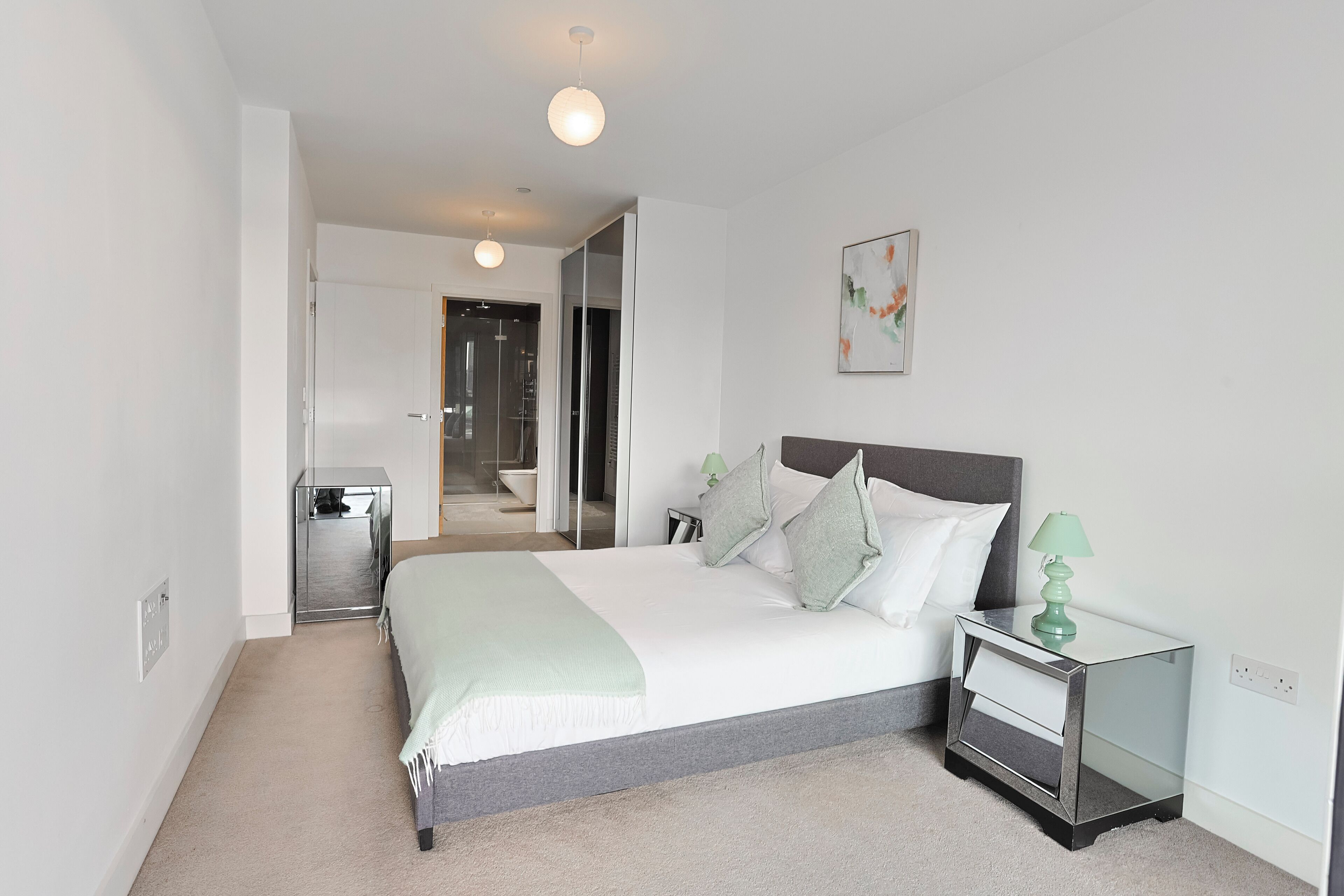 A modern bedroom with a bed, bedside tables, a nightstand, a mirror, and a door leading to a bathroom.