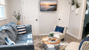 Ein Wohnzimmer mit Sofa, Sessel und Couchtisch, einem an der Wand befestigten Fernseher und einer Tür mit Blick ins Freie.