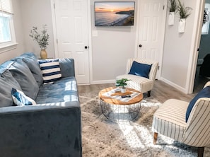 Ein Wohnzimmer mit Sofa, Sessel und Couchtisch, einem an der Wand befestigten Fernseher und einer Tür mit Blick ins Freie.