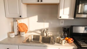 Cocina con gabinetes blancos, un lavamanos de acero inoxidable, una cafetera y un tazón de fruta.