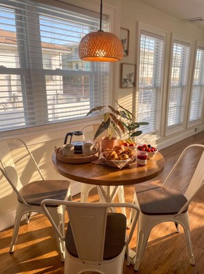 Un comedor con una mesa redonda de madera, sillas blancas, una lámpara colgante de fibra trenzada, una cafetera, pasteles, fruta y una planta.