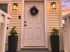 A white front door with a wreath, two potted plants, and two wall-mounted lights. - Unit 1 - Ocean View - Third House from Beach & Boardwalk! - 2BR - Parking (Seaside Heights)