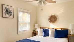 Ein Schlafzimmer mit einem Bett, einem Deckenventilator, einem Fenster mit Jalousien, einer Wandlampe und einem gerahmten Kunstwerk.