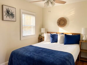Ein Schlafzimmer mit einem Bett, einem Deckenventilator, einem Fenster mit Jalousien, einer Wandlampe und einem gerahmten Kunstwerk.