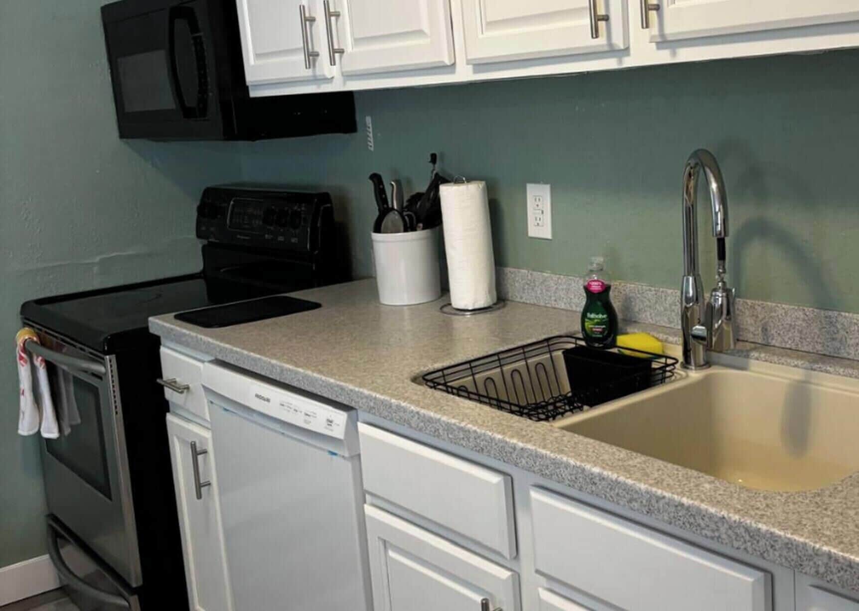 A kitchen with white cabinets, a black microwave, a dishwasher, a sink with a faucet, and a dish rack.