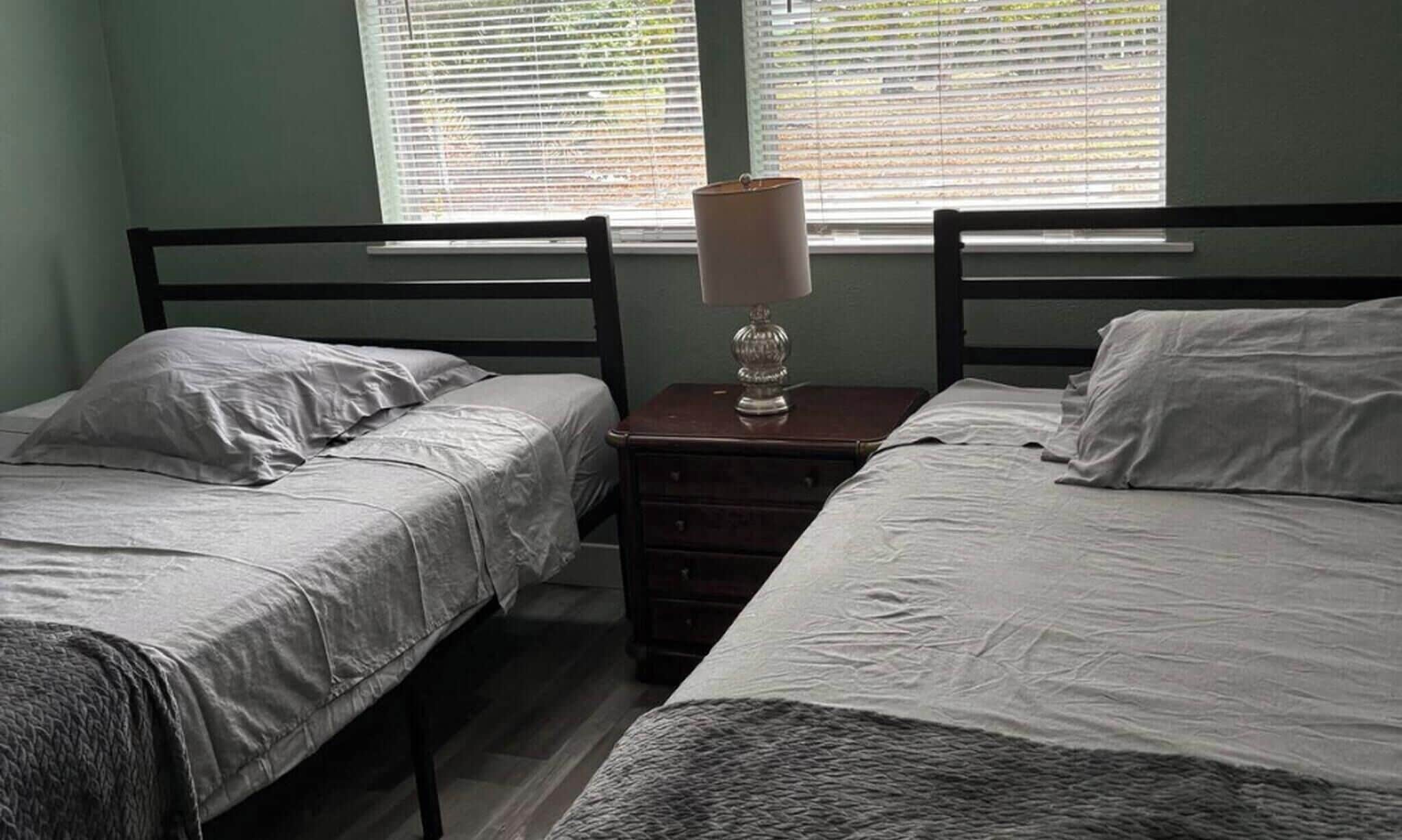 A bedroom with two single beds, a nightstand with a lamp, and a window with blinds.