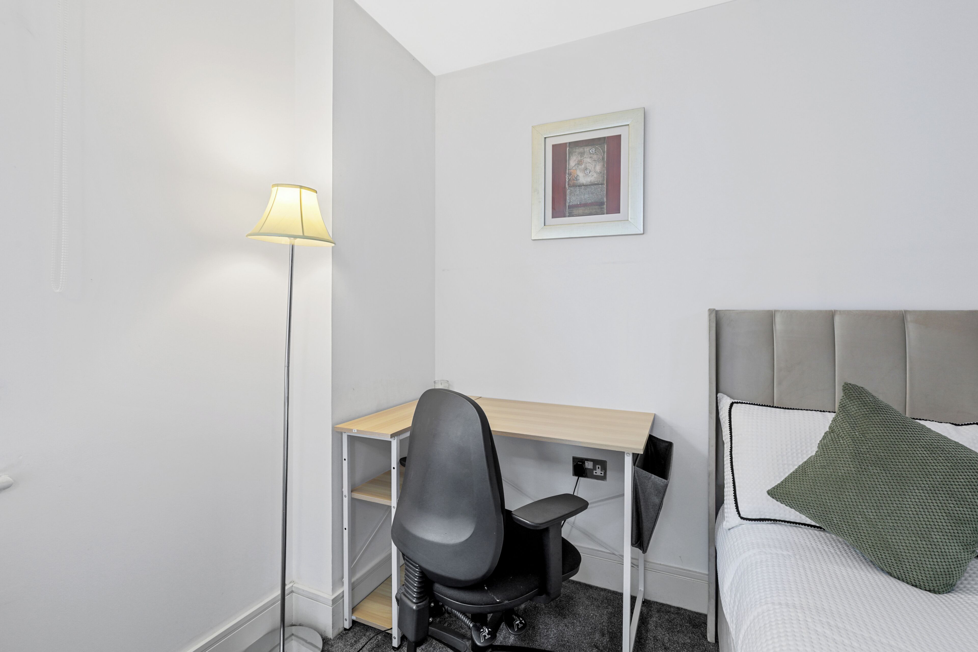 A small, neatly arranged hotel room with a bed, a desk, a chair, and a floor lamp.