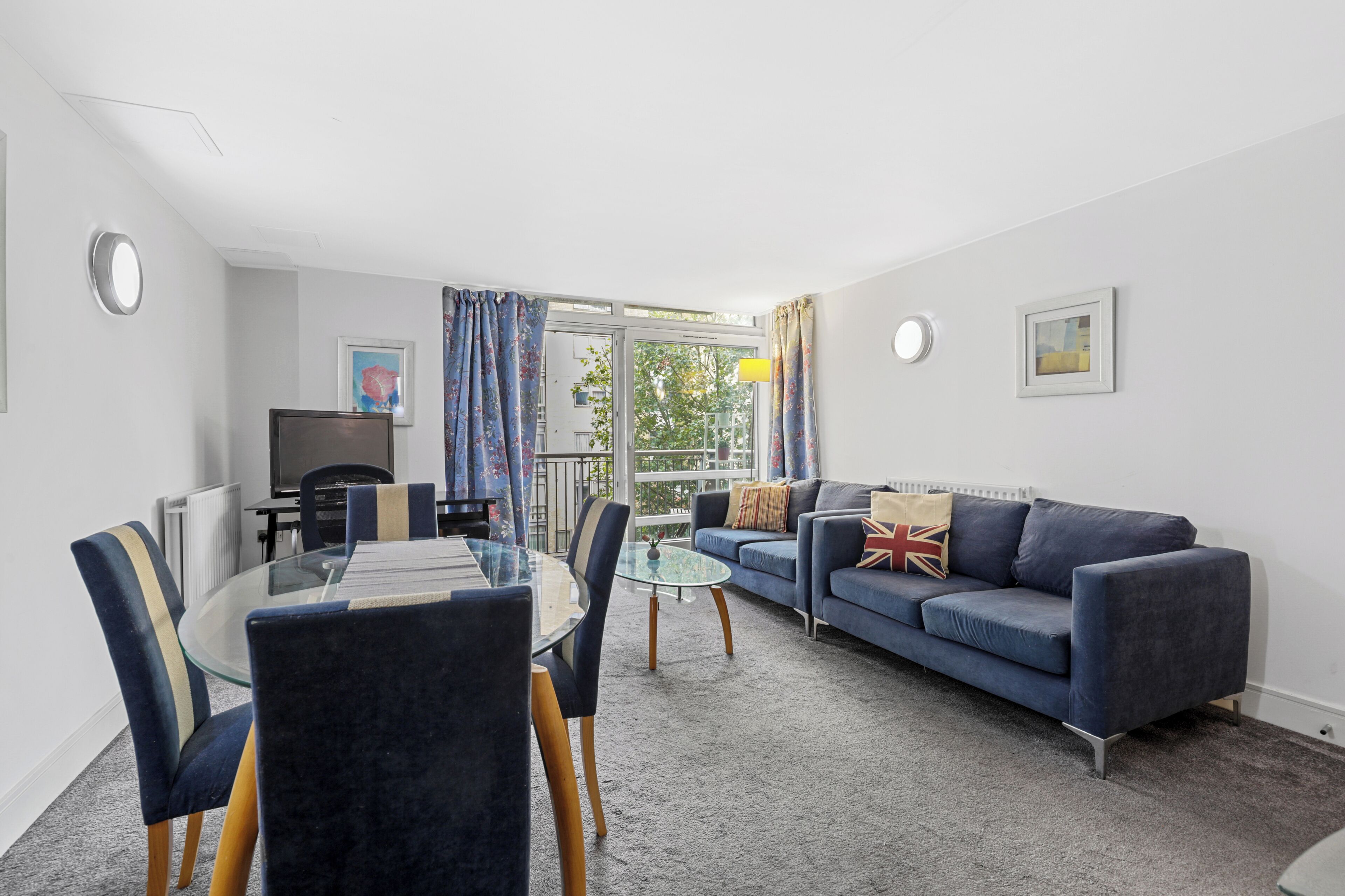 A modern living room with a glass dining table, blue sofa, and a television.