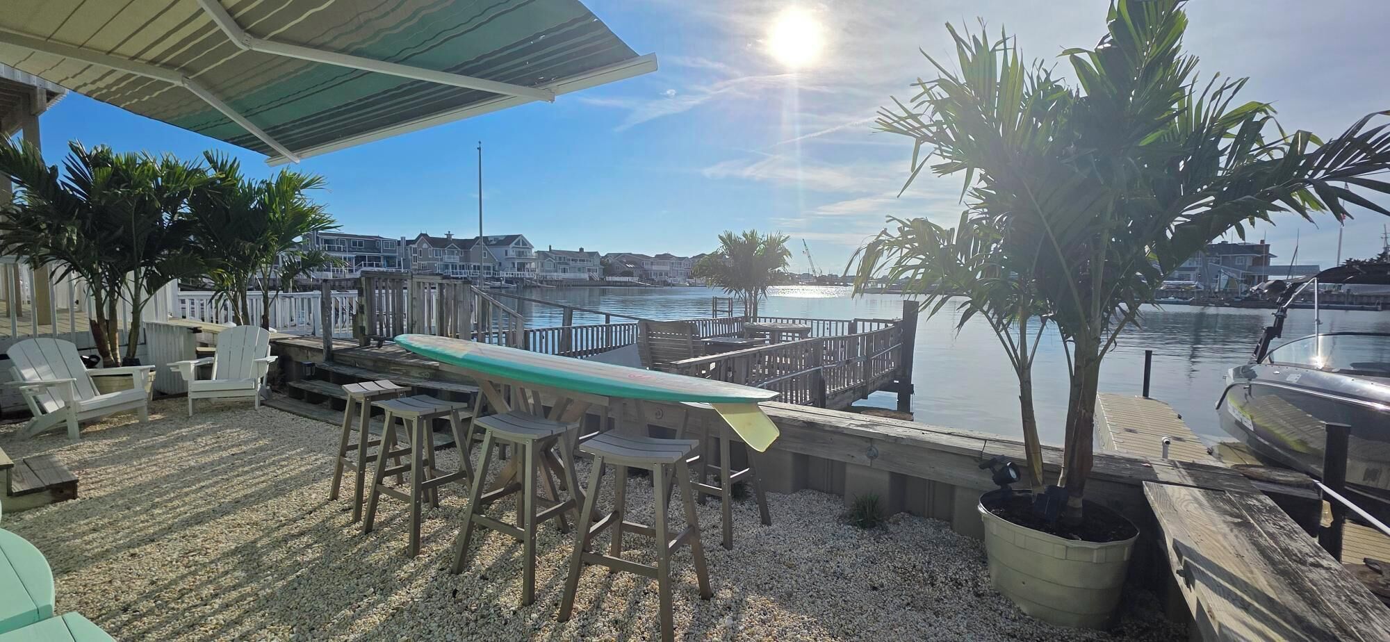 Un patio frente al agua con una mesa larga y sillas, una tabla de surf y vistas al puerto deportivo y al agua.