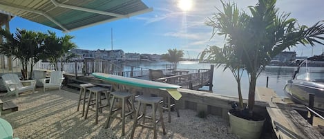 Un patio frente al agua con una mesa larga y sillas, una tabla de surf y vistas al puerto deportivo y al agua.